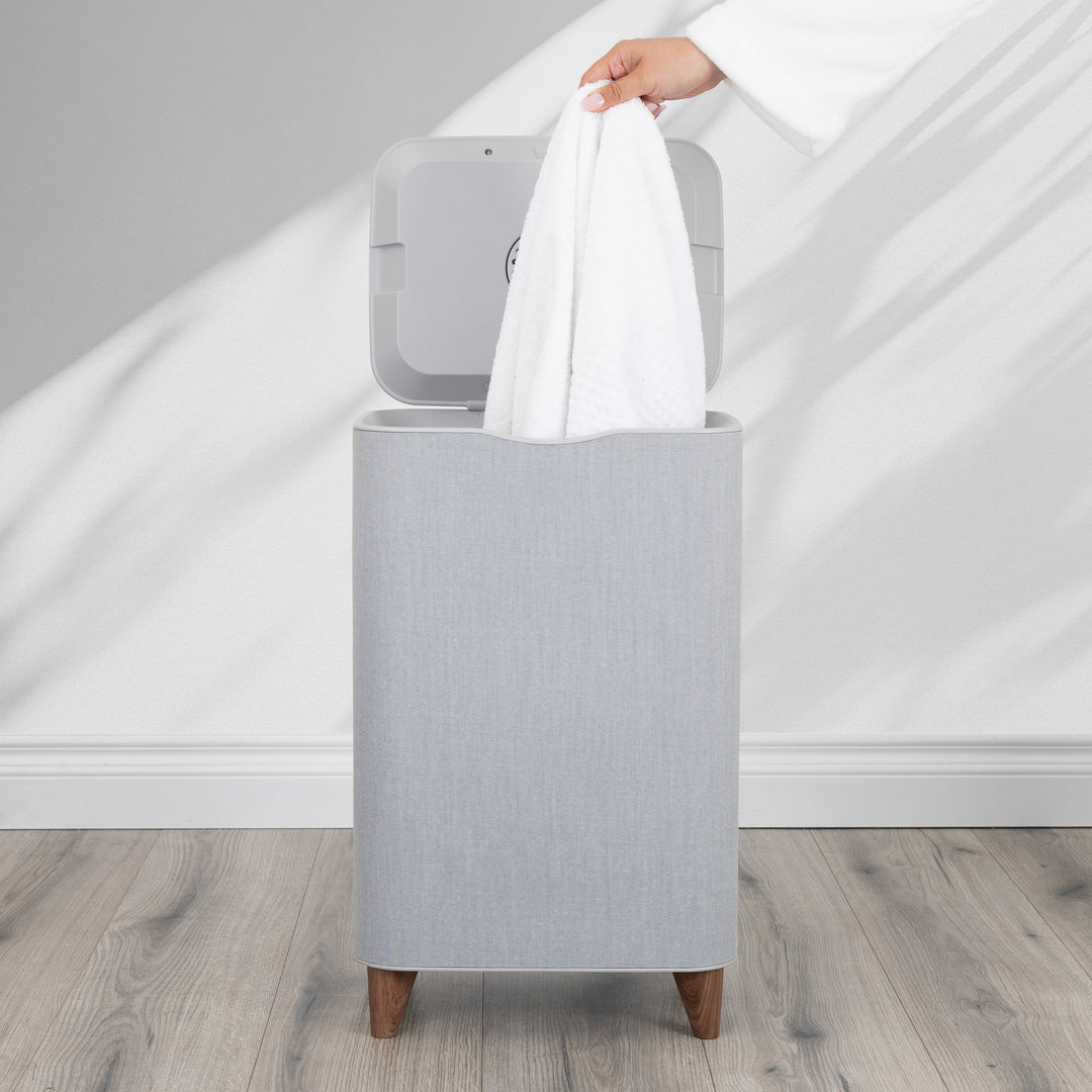 Gray towel warmer basket with a white towel being taken out against a neutral background and wooden bathroom floor