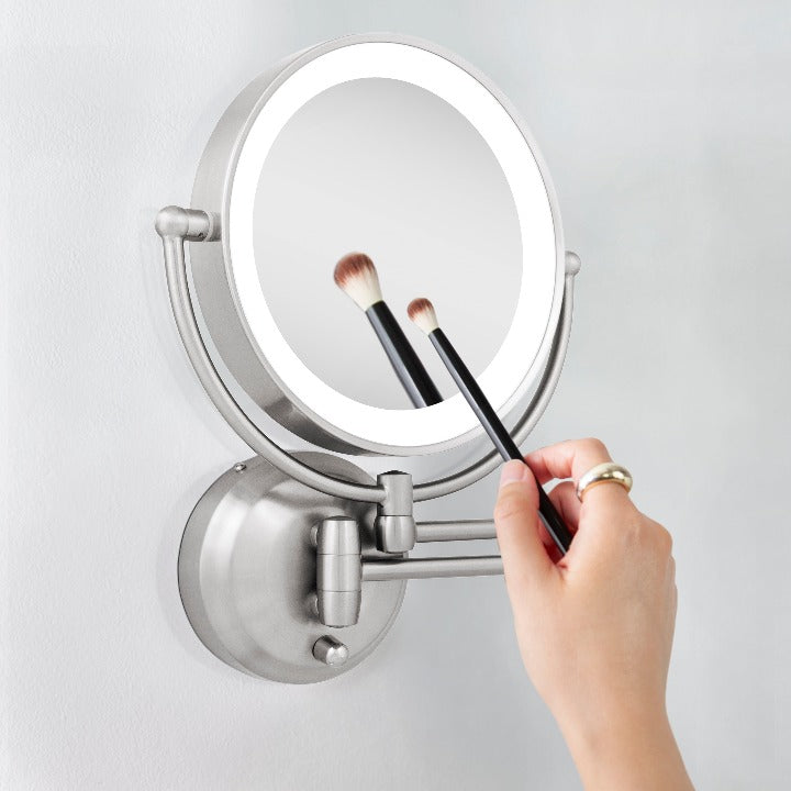 Zadro LEDW410 705004419475 in-use photo side view hand holding brush to magnified mirror , lighted wall mounted makeup mirror with magnification & extendable arm in a real life setting
